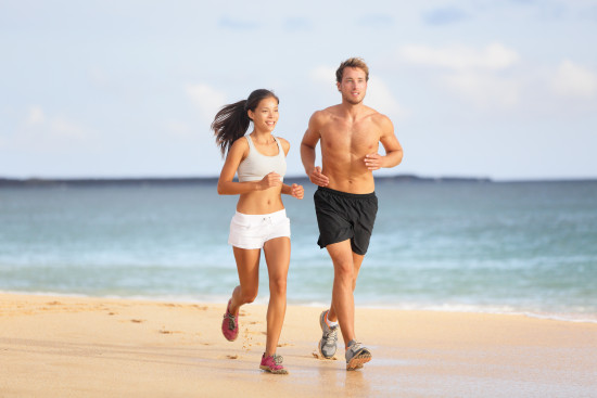 Människor som springer - unga par joggar på stranden. Attraktiv passande sportiga unga parlöpare sida vid sida på stranden i sommarsolen njuter av den friska luften när de tränar tillsammans